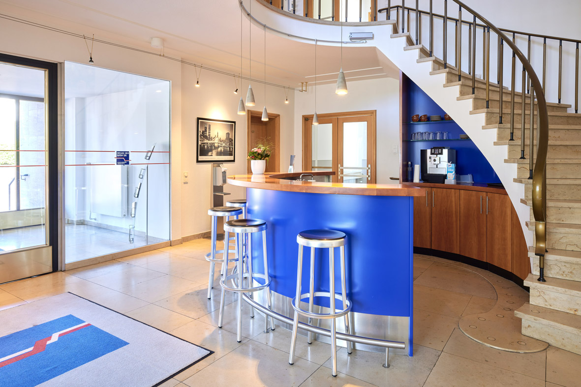 Moderner Eingangsbereich mit blauem Empfangstresen und drei hohen Metallhockern des Remscheider Standorts von Kaib, Galldiks und Partner.