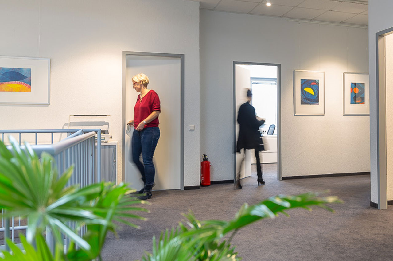 Büroflur mit zwei Personen, mehreren farbigen abstrakten Bildern an der Wand und grünen Pflanzen im Vordergrund.