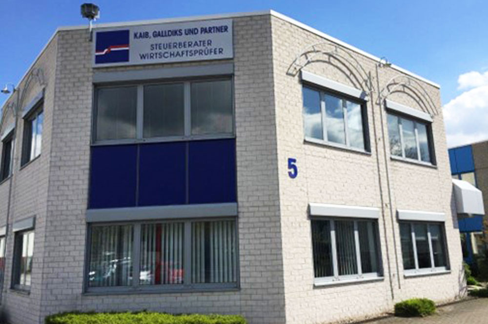 Das zweigeschossige Bürogebäude in Hilden mit hellen Fassadensteinen, blauen Fensterrahmen und Firmenbeschriftung von Kaib, Galldiks und Partner.